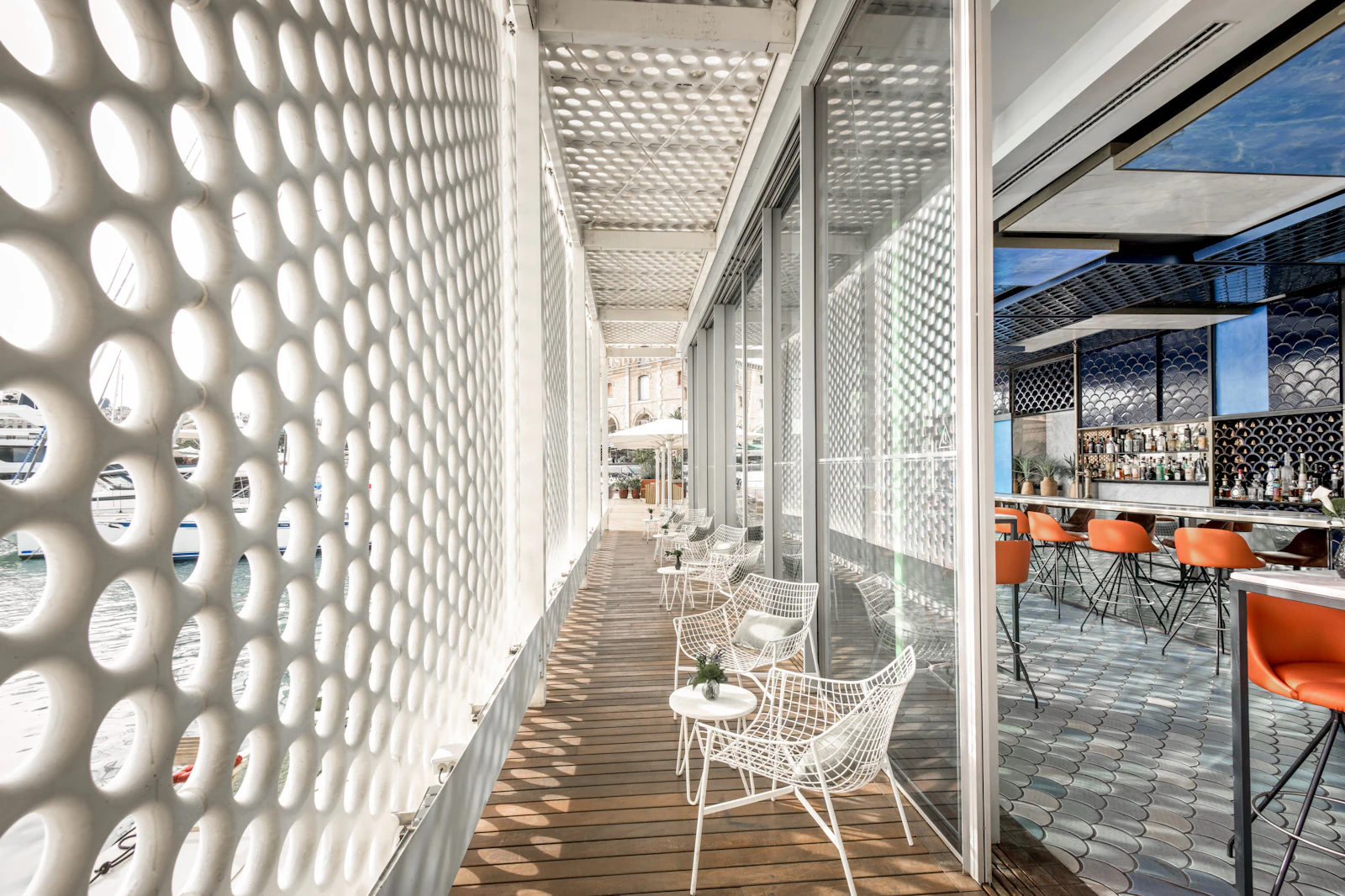 Covered waterfront terrace with white perforated screens, wooden deck, white chairs, and bar area with orange seating visible through glass doors.