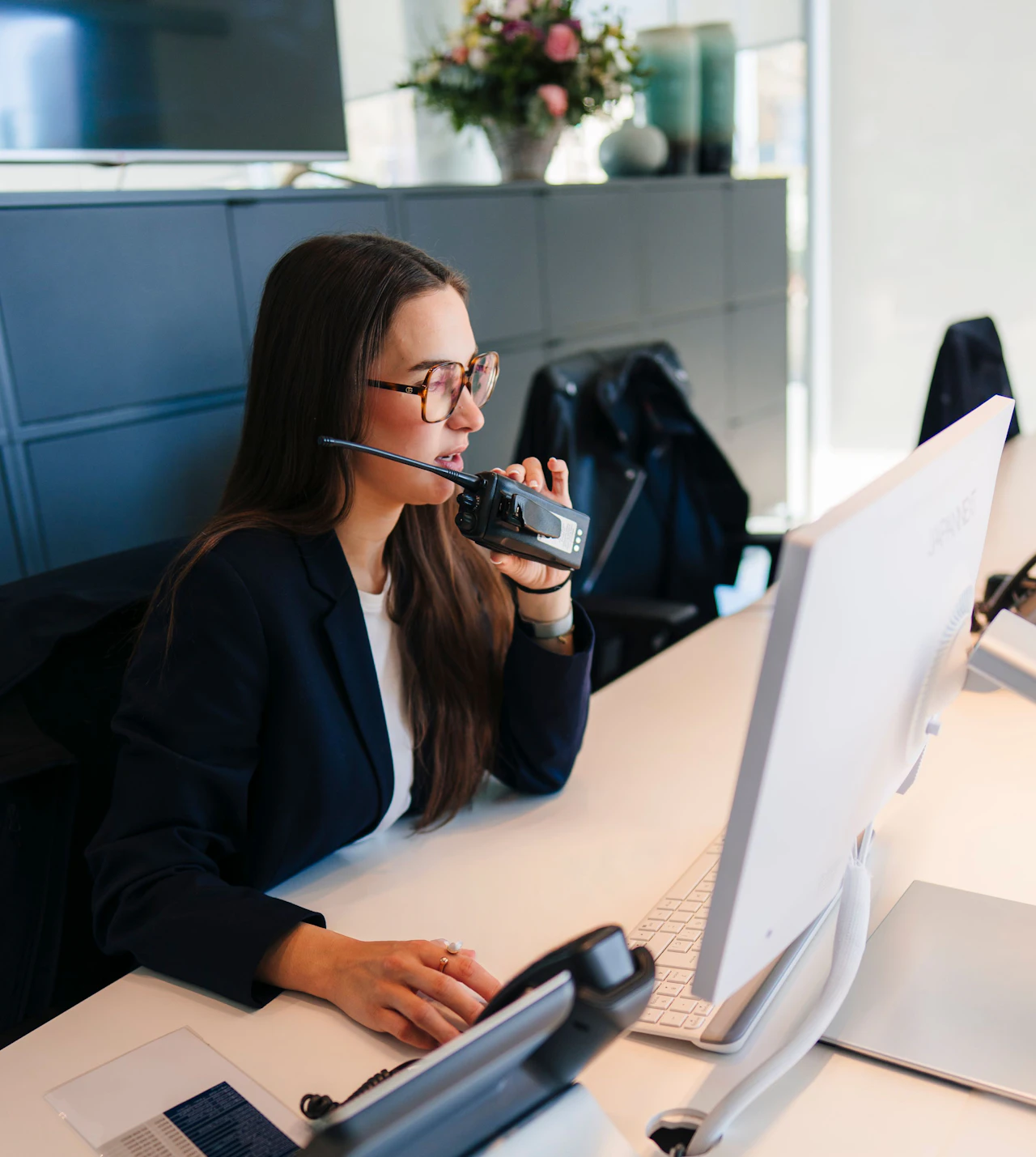 Woman wearing glasses and headset speaking into radio handset at office desk with computer.