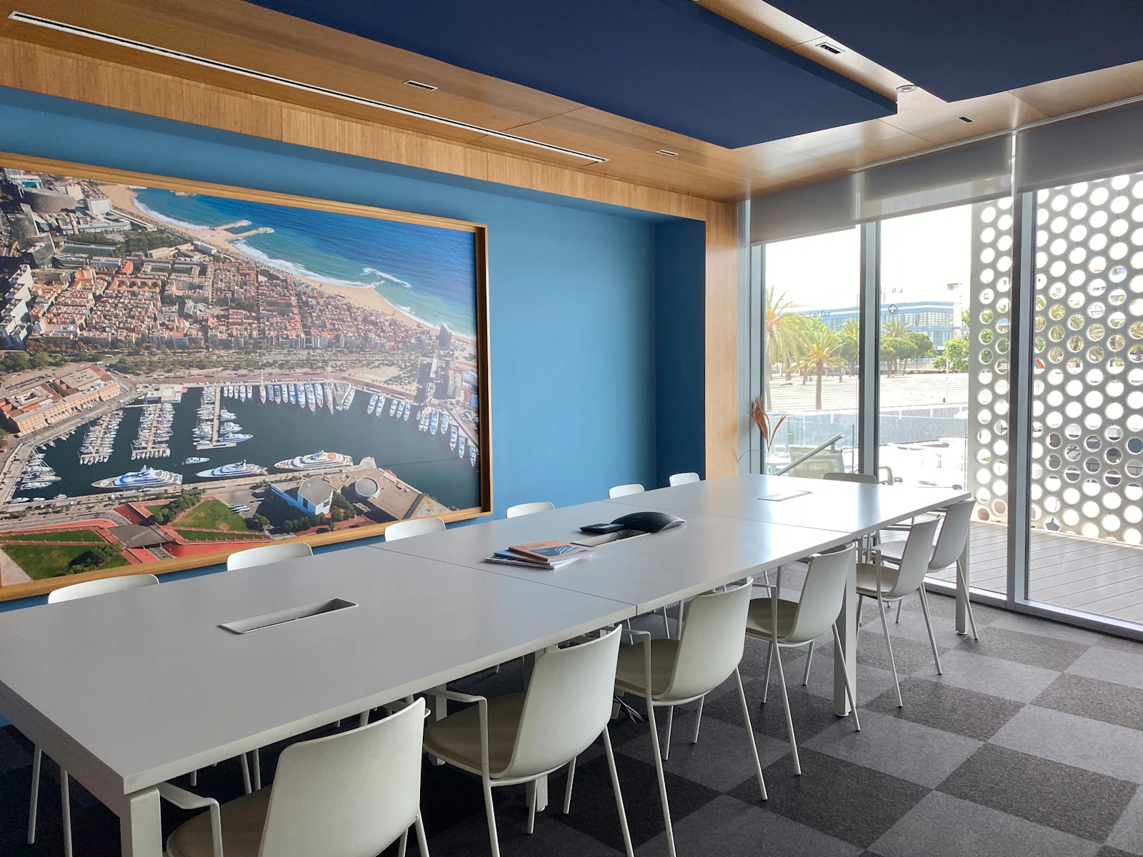 Sala de conferencias moderna con una fotografía aérea del puerto deportivo en la pared azul, con vistas al litoral de Barcelona a través de puertas de cristal.