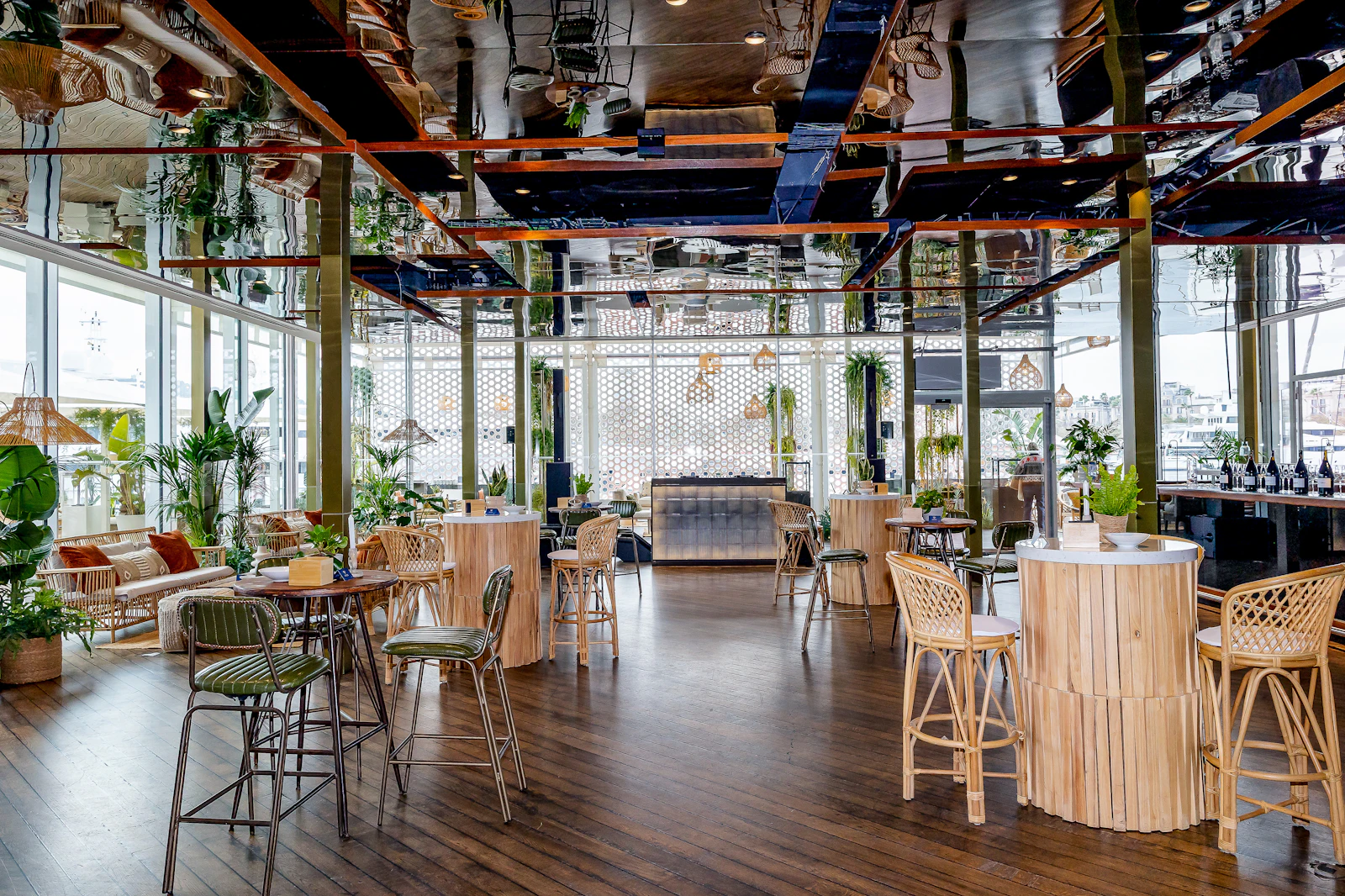 Restaurant interior with high wooden tables, rattan stools, plants and a mirrored ceiling.