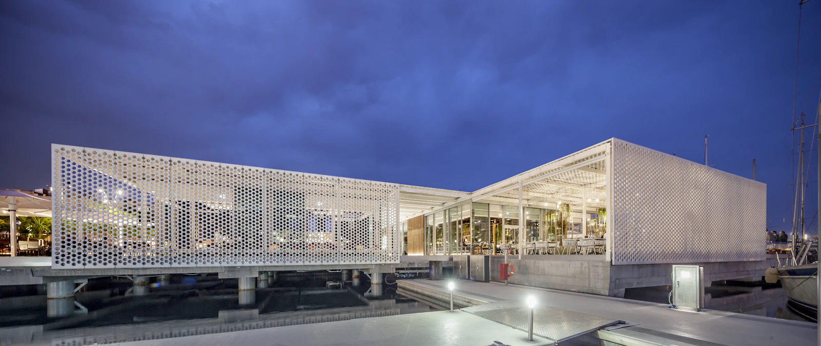 Illuminated modern waterfront building with perforated white facade at dusk beside moored boats.