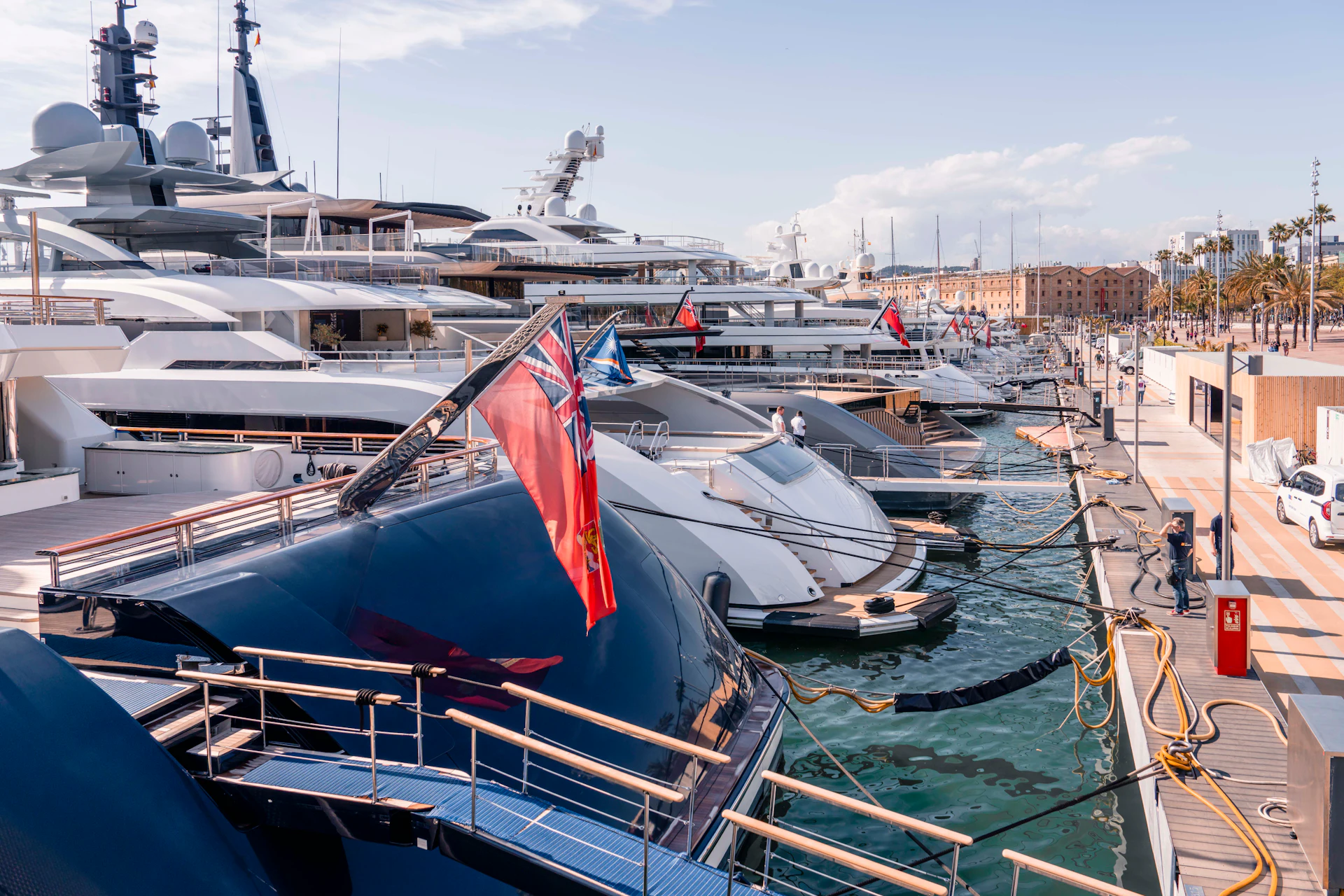Multiple superyachts moored at Port Vell marina in Barcelona with waterfront buildings and palm trees visible.