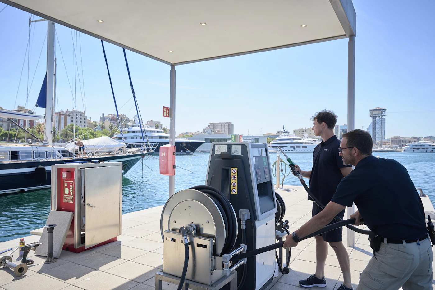 Two men handle a fuel pump and hose at a marina fuelling station with yachts behind.