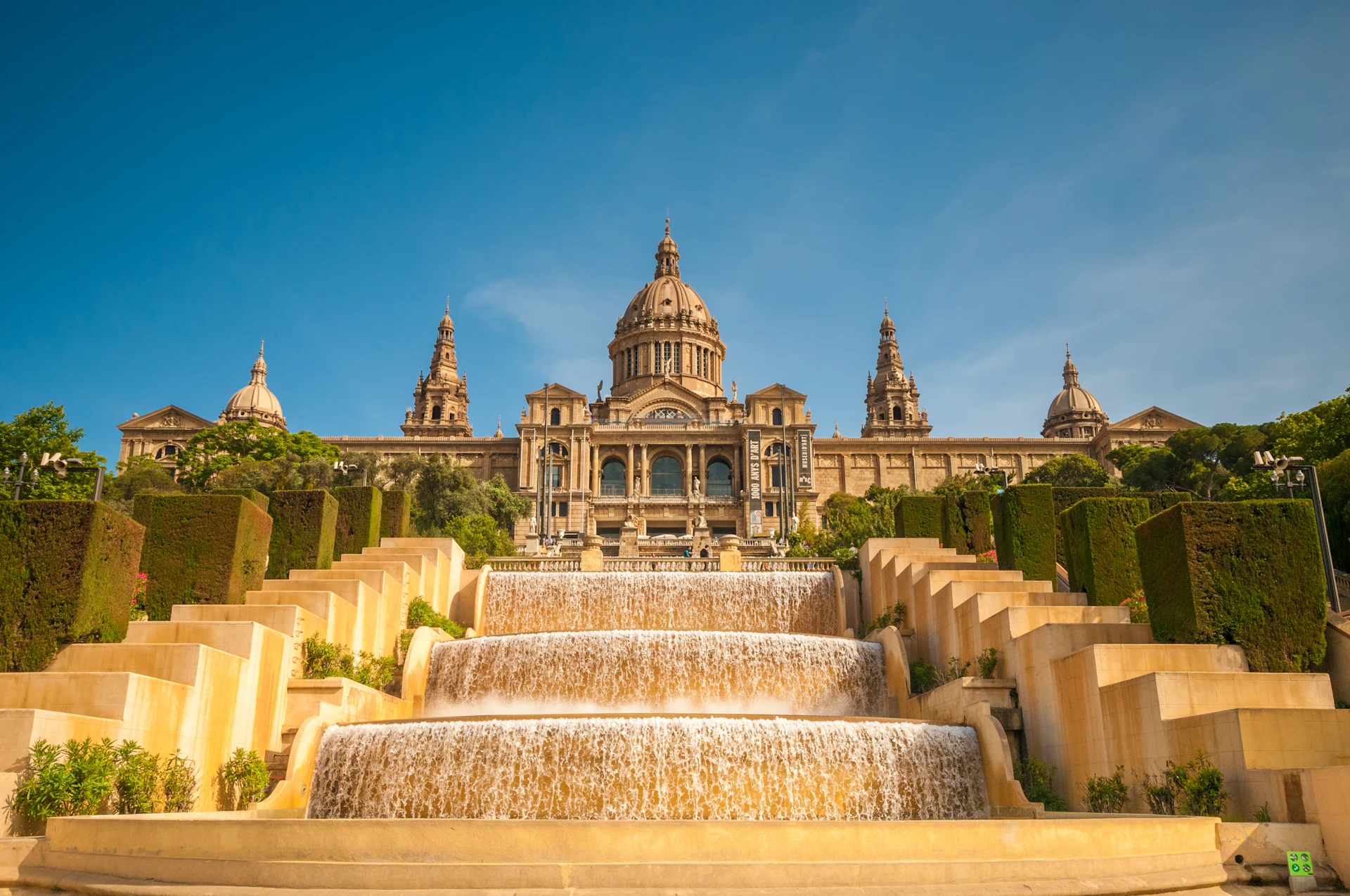 Cascading fountains with manicured hedges leading to a grand domed palace under clear blue sky.