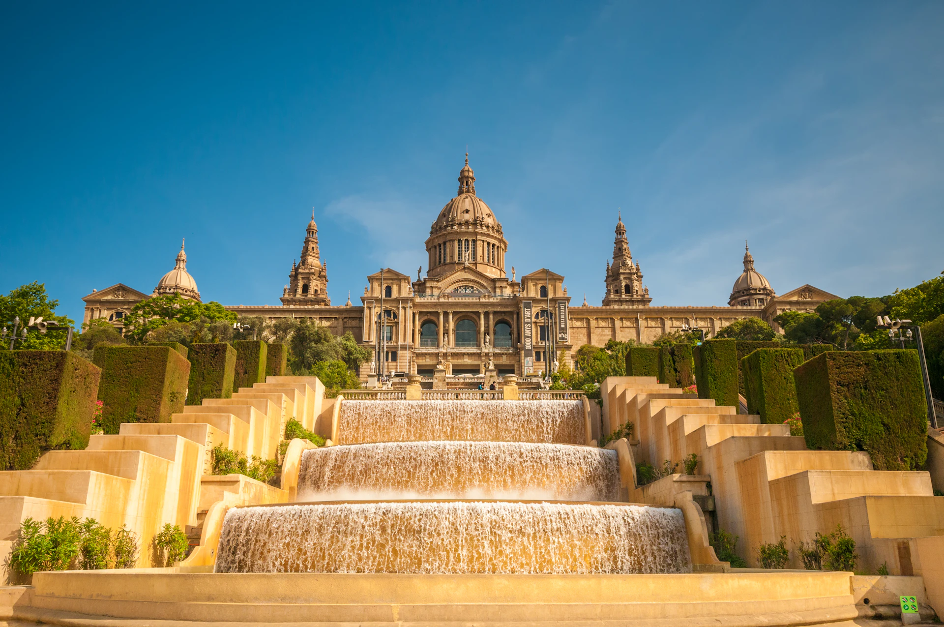 Cascading fountains with manicured hedges leading to a grand domed palace under clear blue sky.