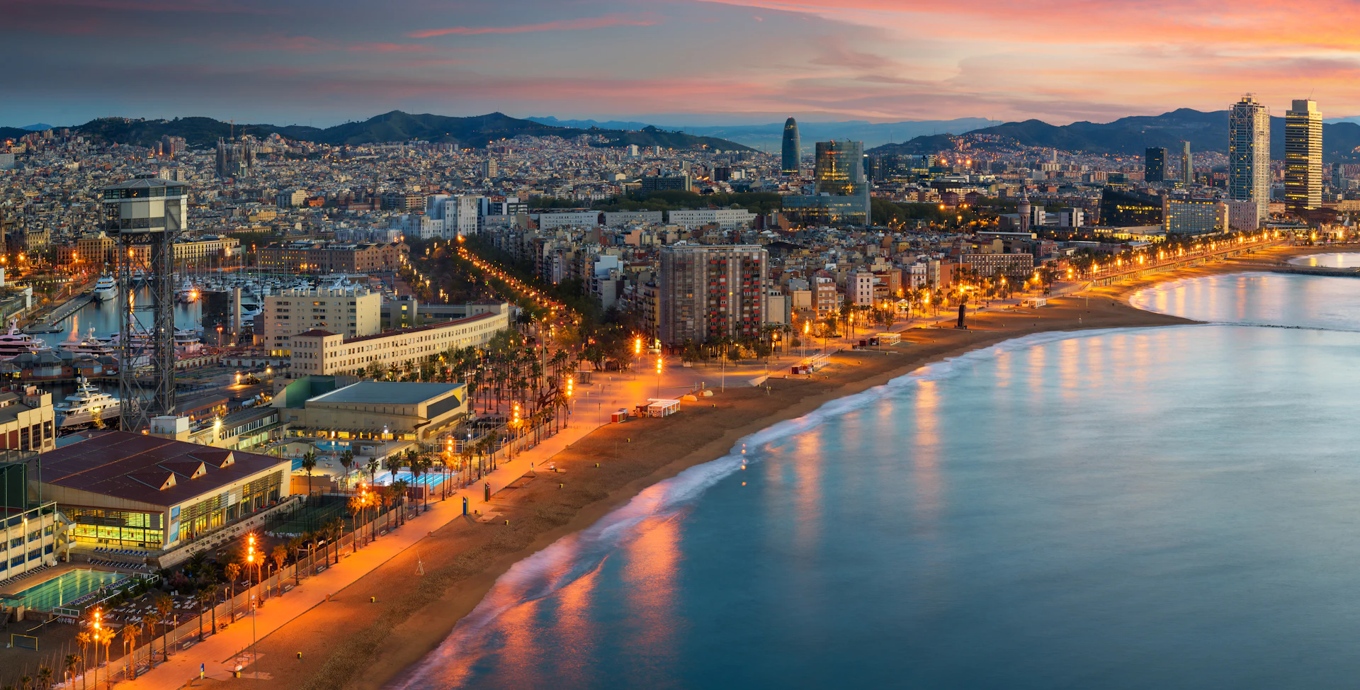 Aerial view of Barcelona's illuminated beachfront and city skyline at dusk.