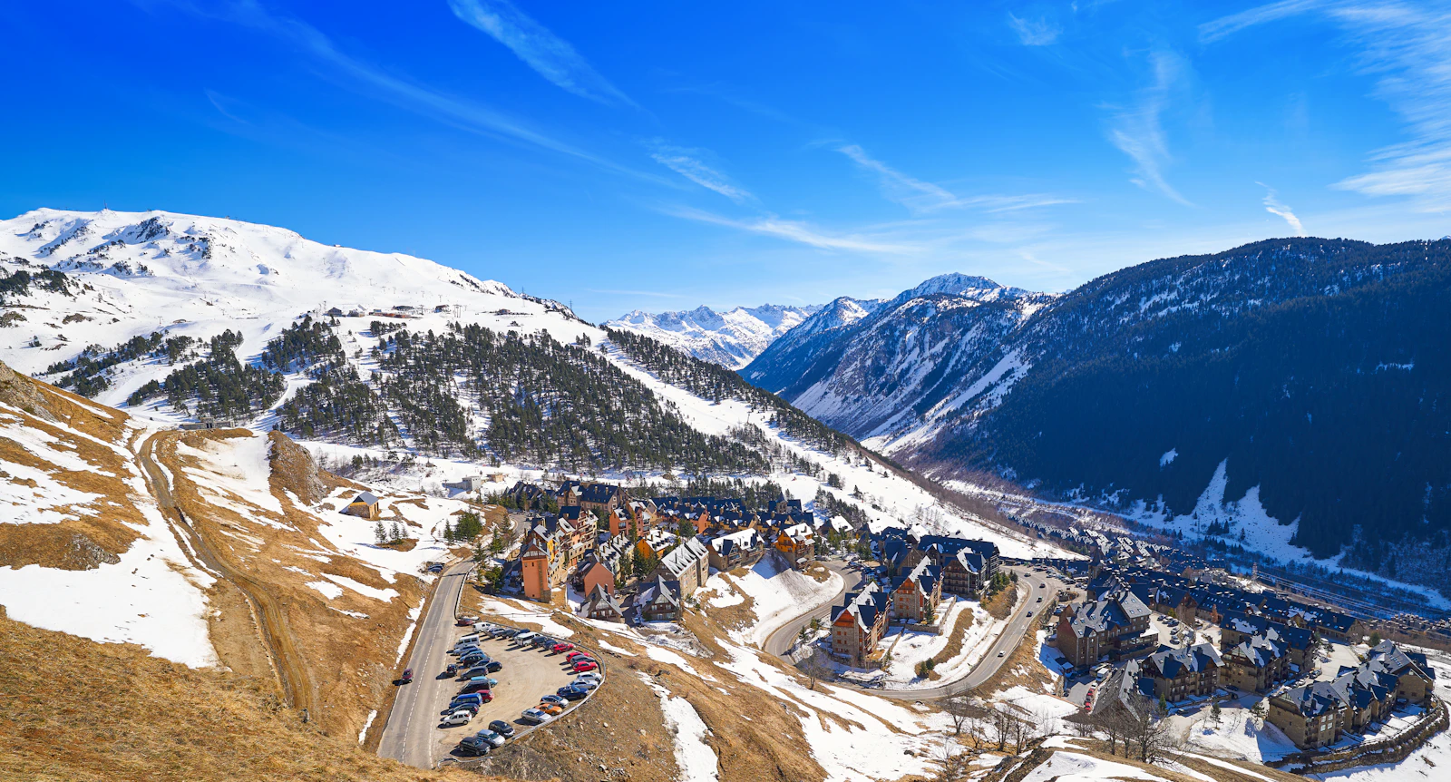 Snowy mountain valley with a ski resort village and winding roads below blue skies.