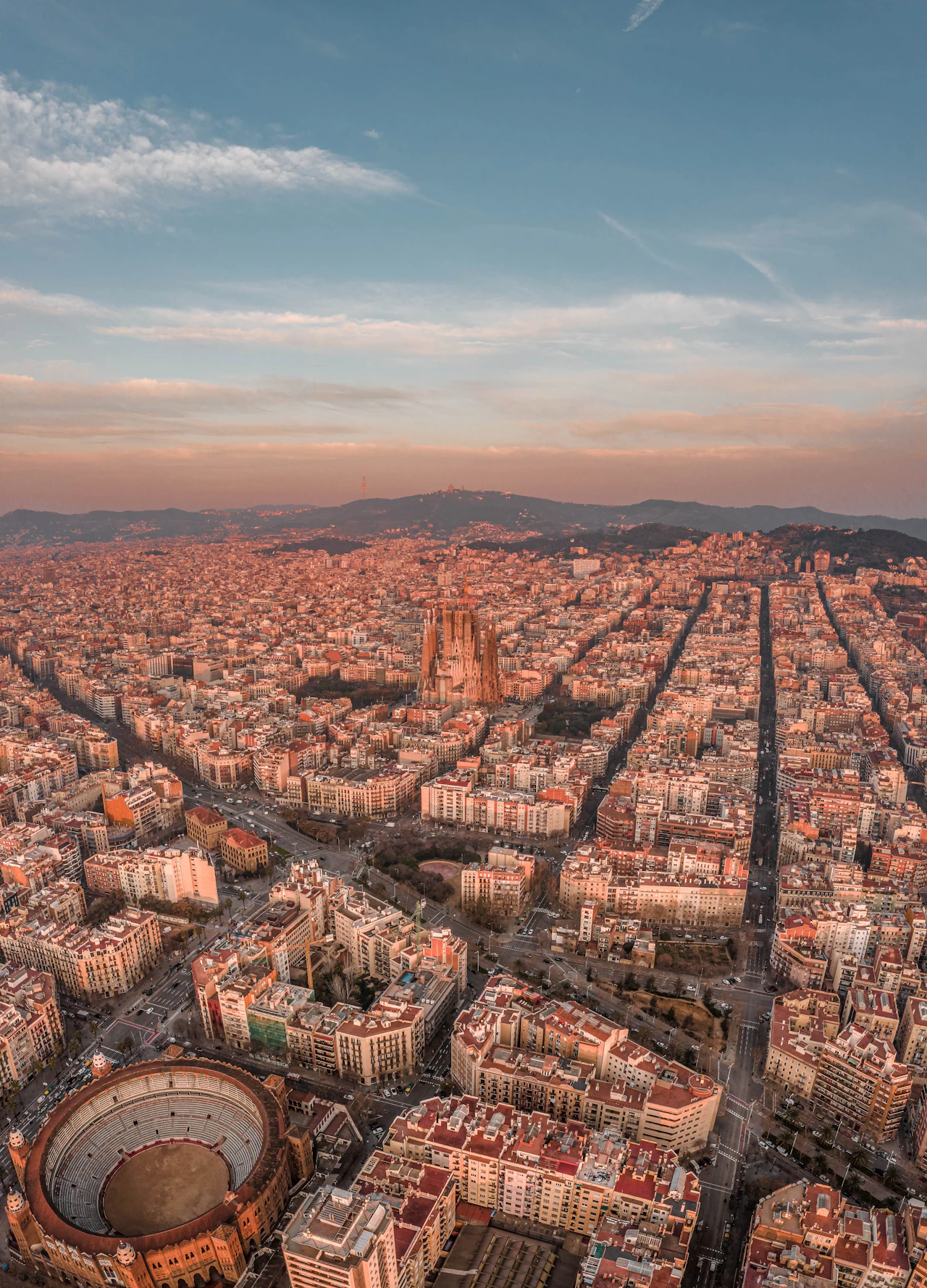 Aerial view of Barcelona at sunset showing the cityscape, Sagrada Familia basilica, and surrounding hills.