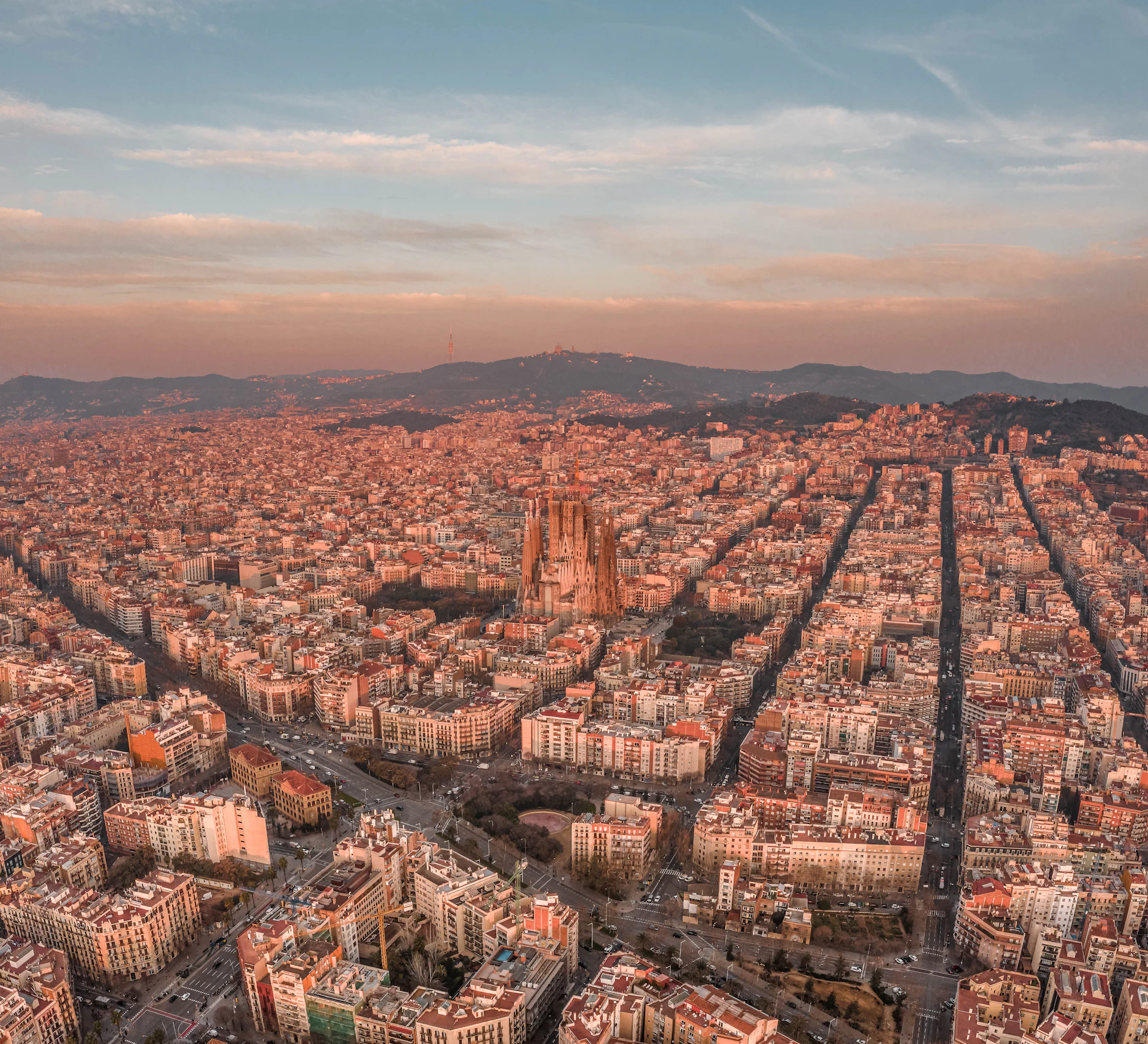 Aerial view of Barcelona at sunset with the Sagrada Família and hills beyond.