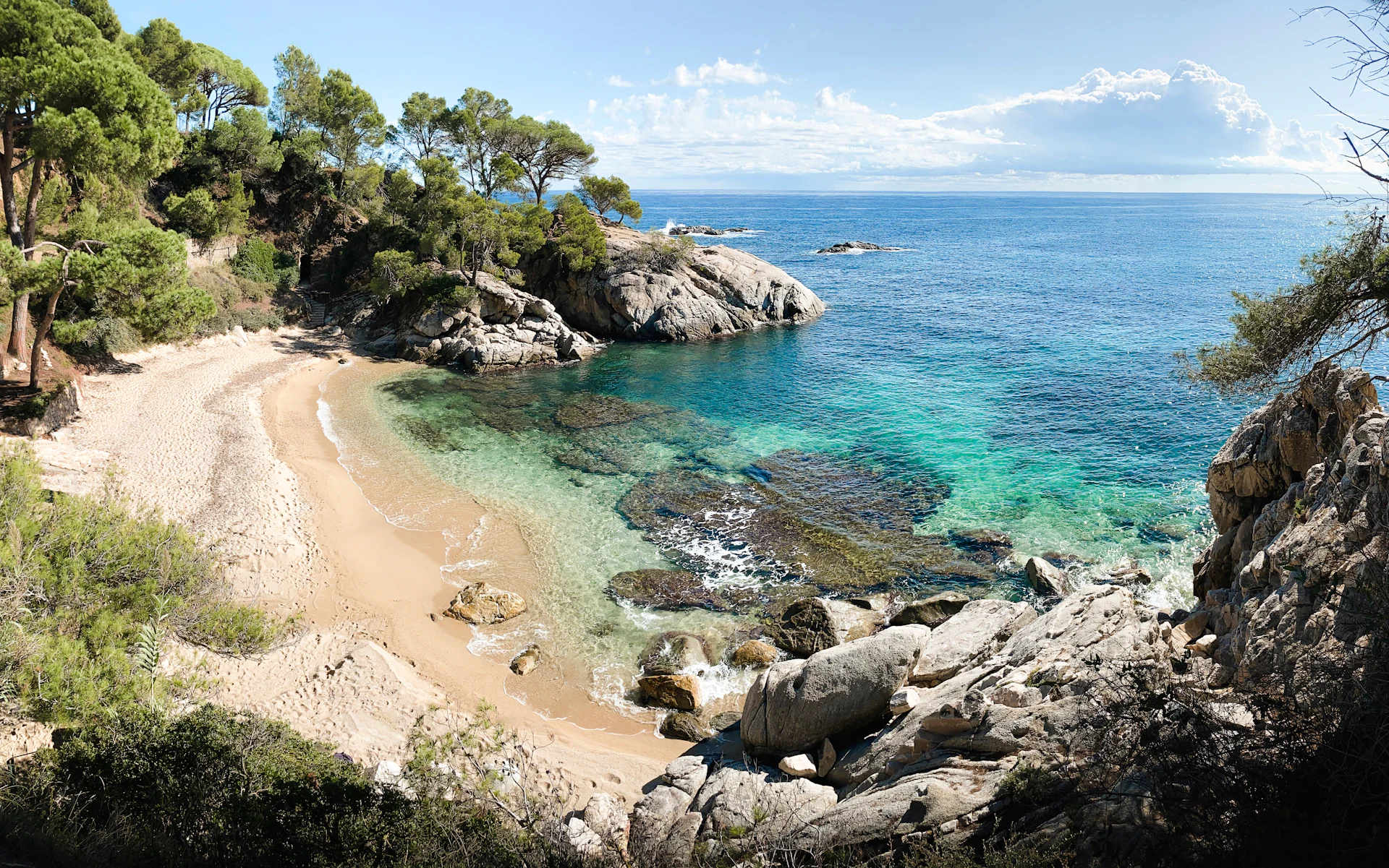 Secluded sandy cove with turquoise water, rocky outcrops and pine trees along the coast.