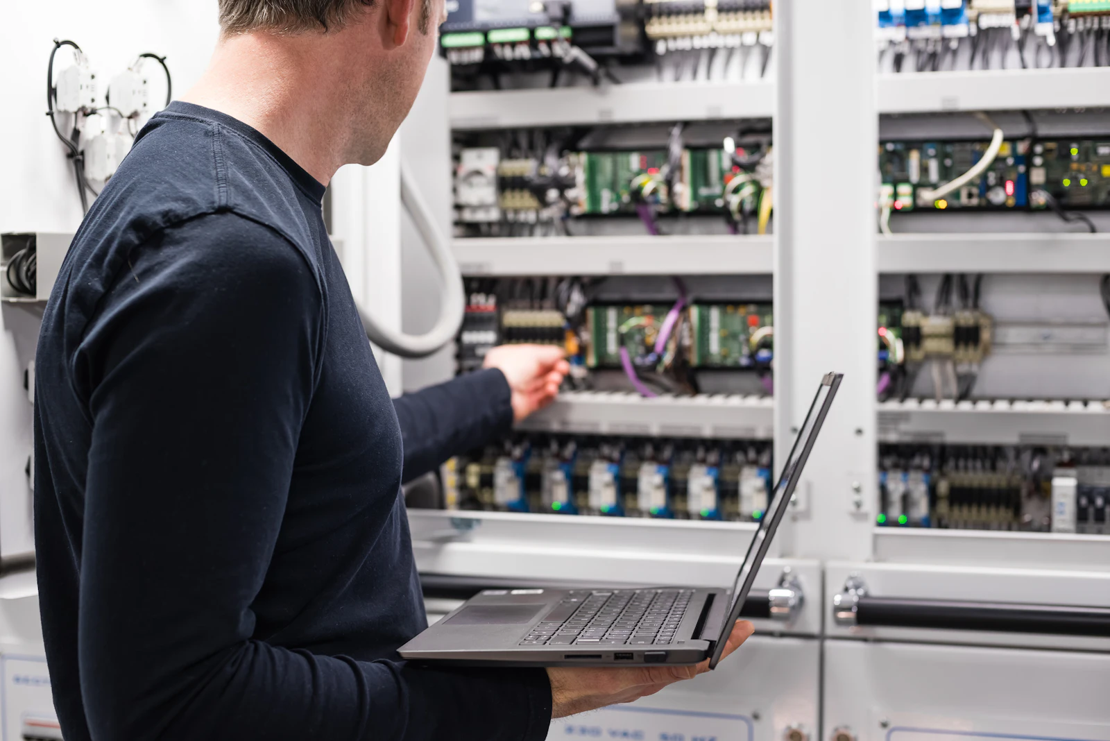 Técnico sosteniendo un portátil mientras inspecciona un cuadro de control eléctrico con placas de circuitos y cableado.