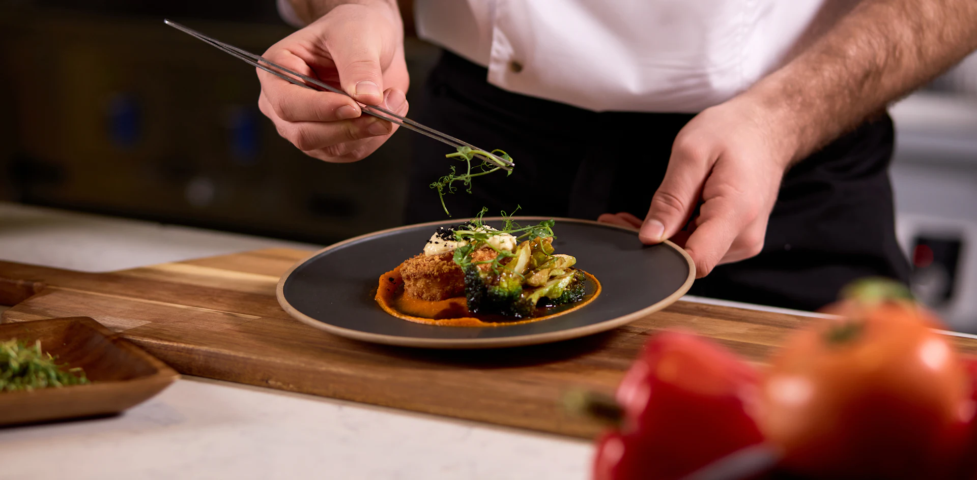 Chef plating a fine dining dish with tweezers, garnishing with microgreens over an orange sauce.