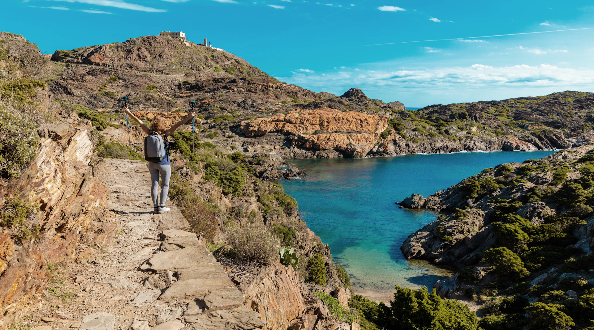 Hiker with backpack walking along a coastal cliff path above turquoise Mediterranean water.