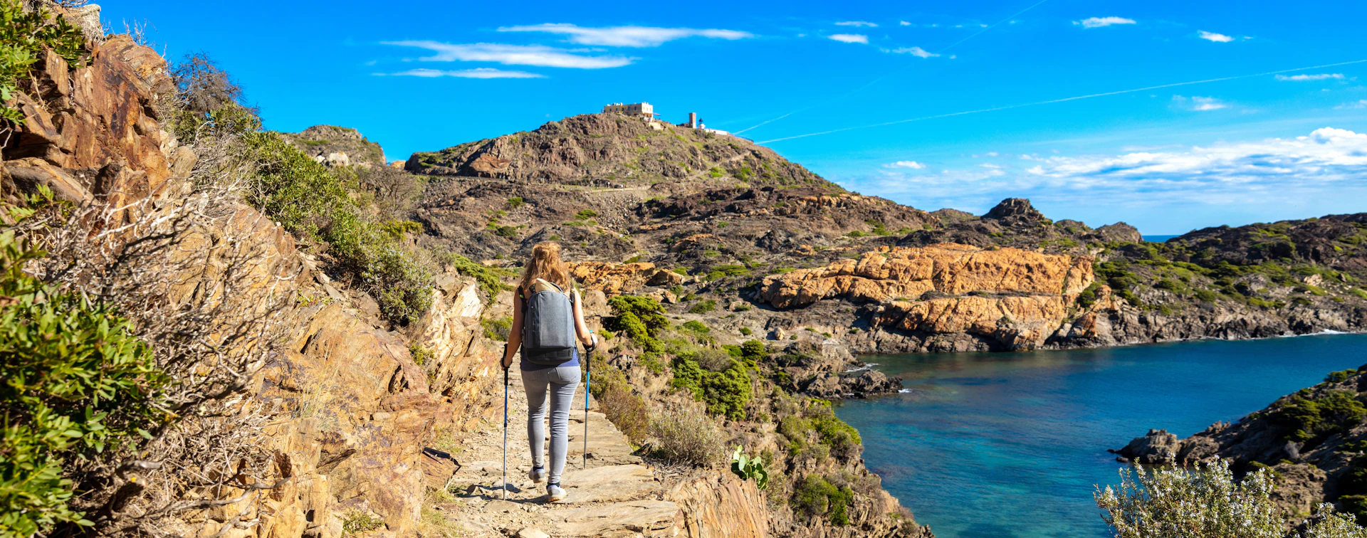 Hiker with backpack walking on coastal cliff path above blue sea and rocky landscape.