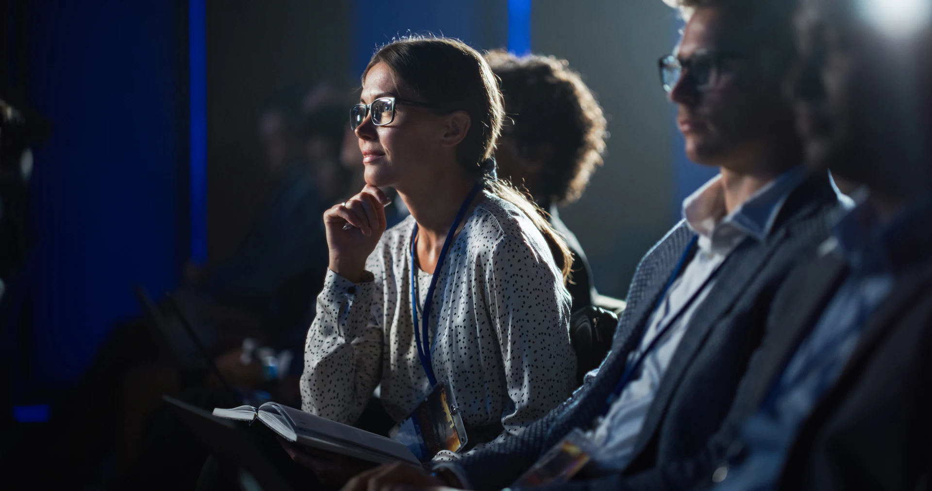 Woman wearing glasses and lanyard sits among business professionals at an indoor event.