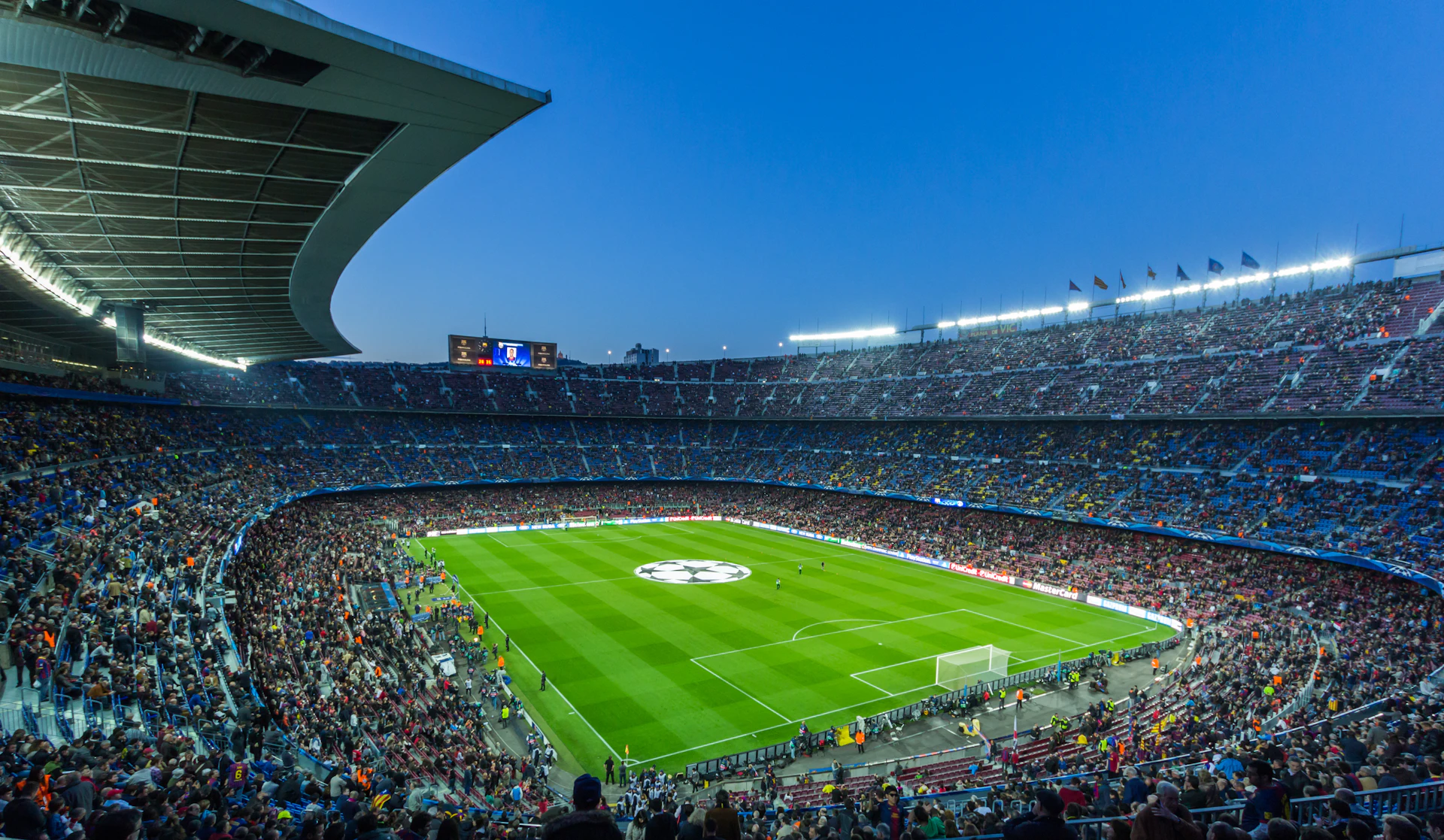 Packed football stadium at dusk with crowds surrounding the illuminated green pitch.