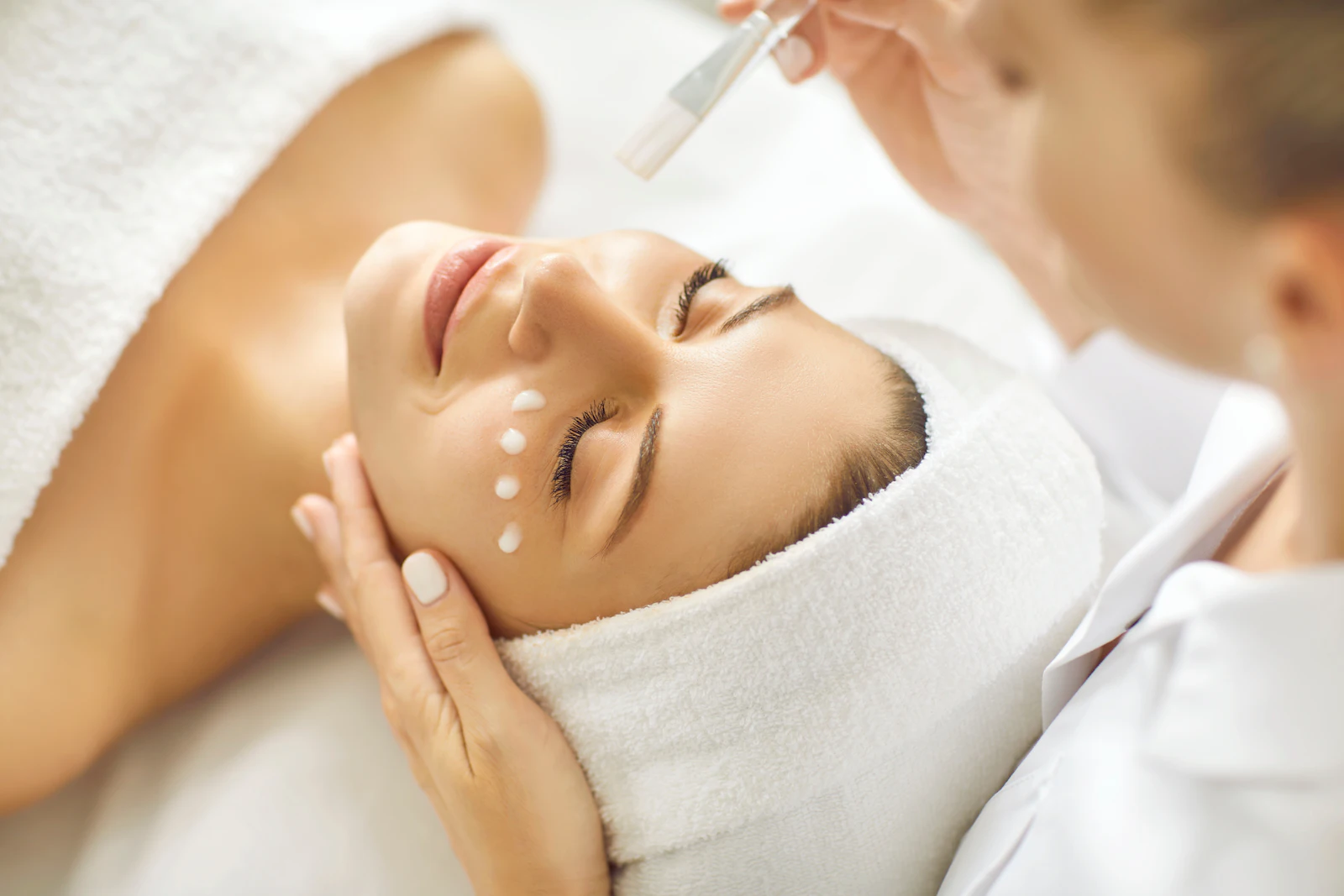 Therapist applies cream dots to a relaxed woman's face during a facial treatment.
