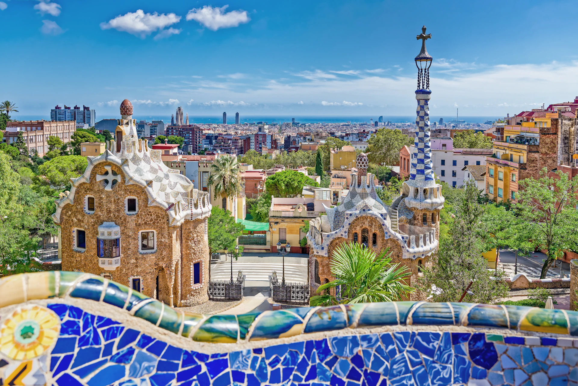 Colorful mosaic railing overlooks ornate gatehouses and Barcelona skyline with Mediterranean Sea.