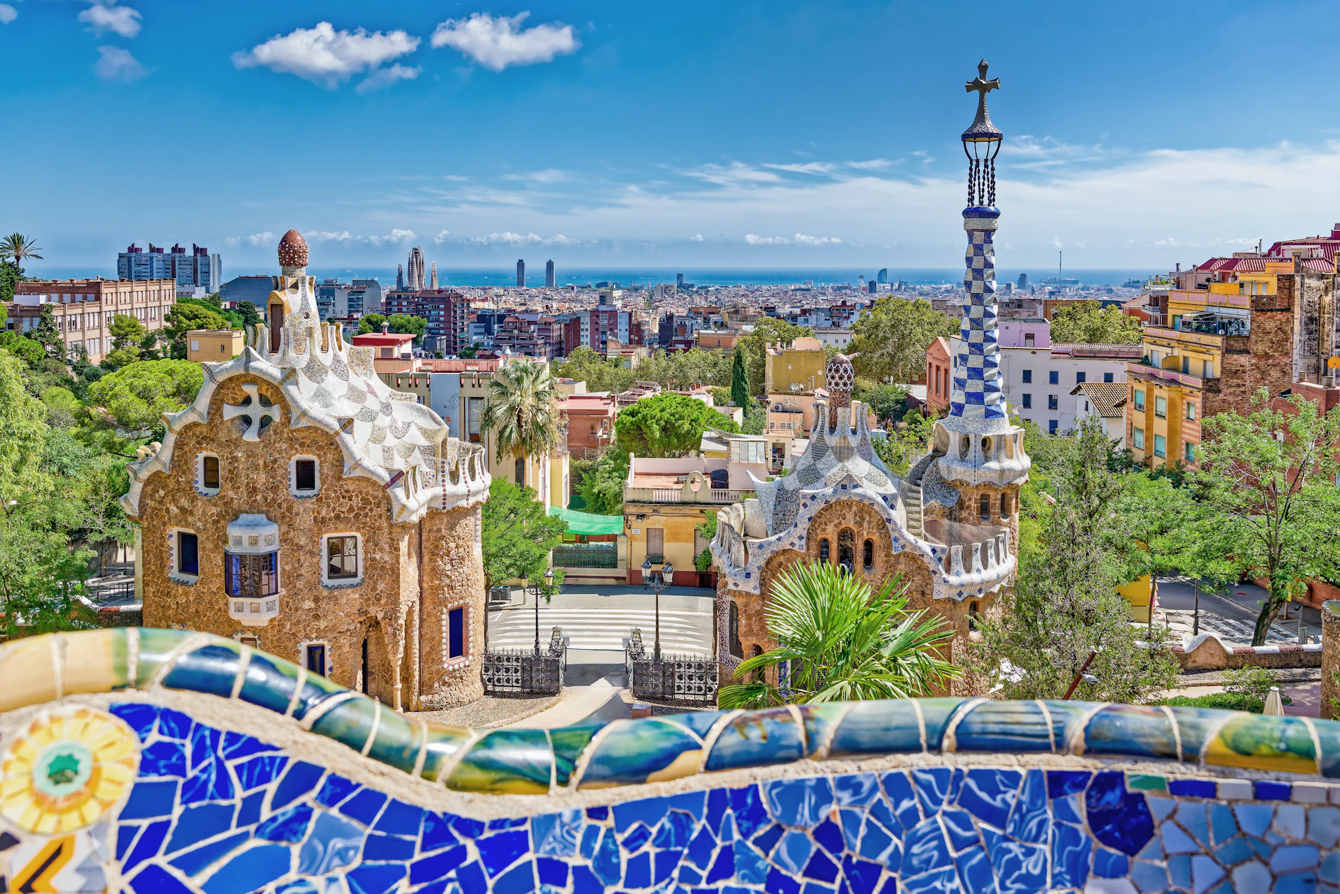 Colorful mosaic railing overlooks ornate gatehouses and Barcelona skyline with Mediterranean Sea.