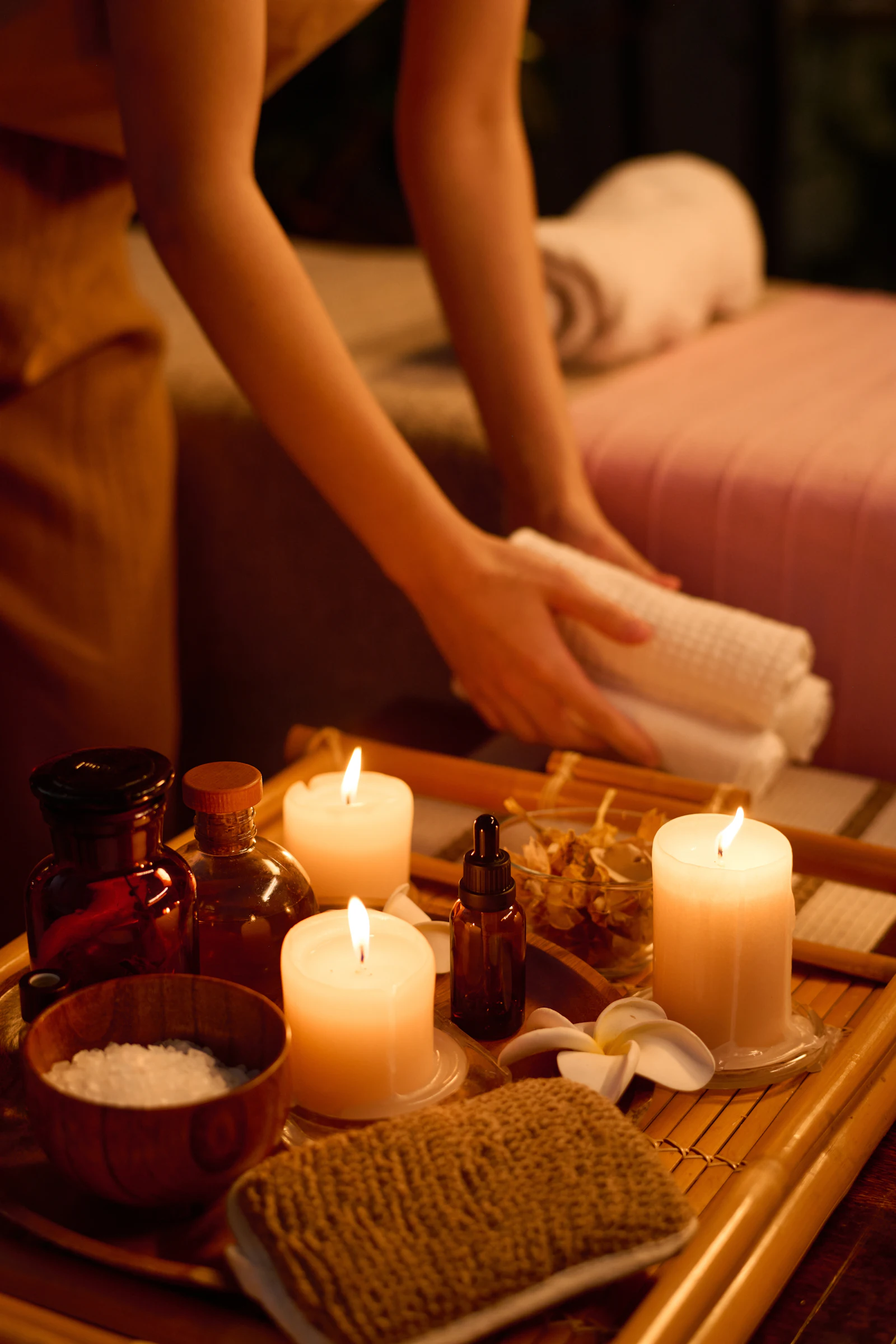 Spa treatment setup with lit candles, essential oils, salt bowl, and therapist's hands holding rolled towels.