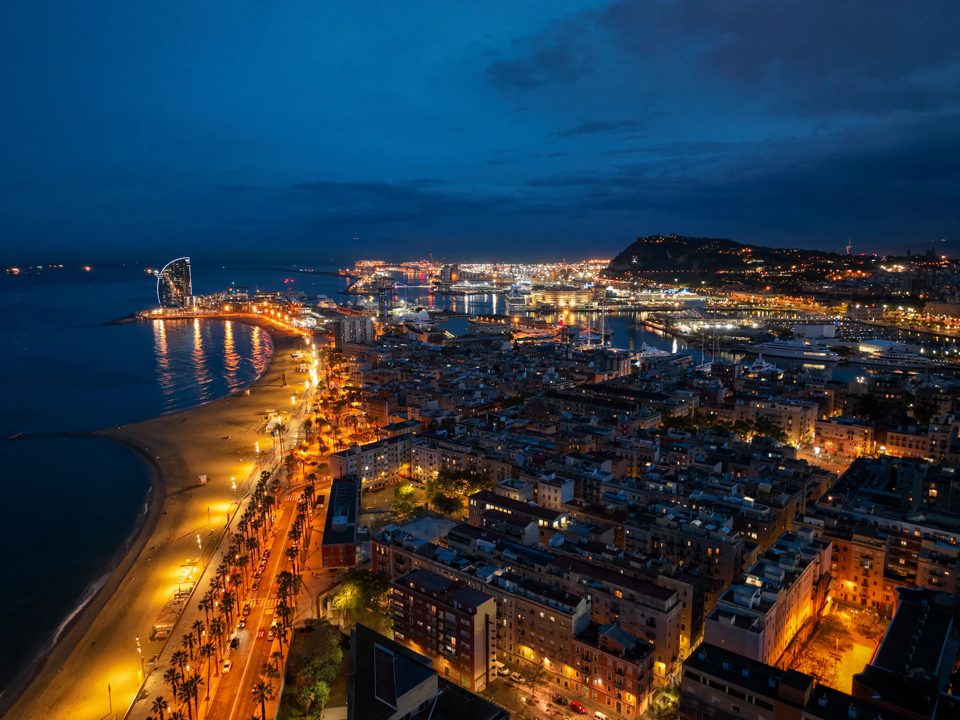 Barcelona coastline at dusk with illuminated beach, buildings, and harbor extending to distant hills.