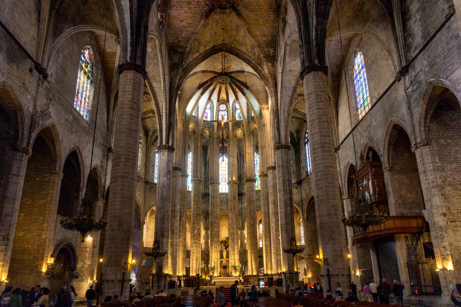 Gothic church interior with tall stone columns, stained glass windows and visitors near the altar.