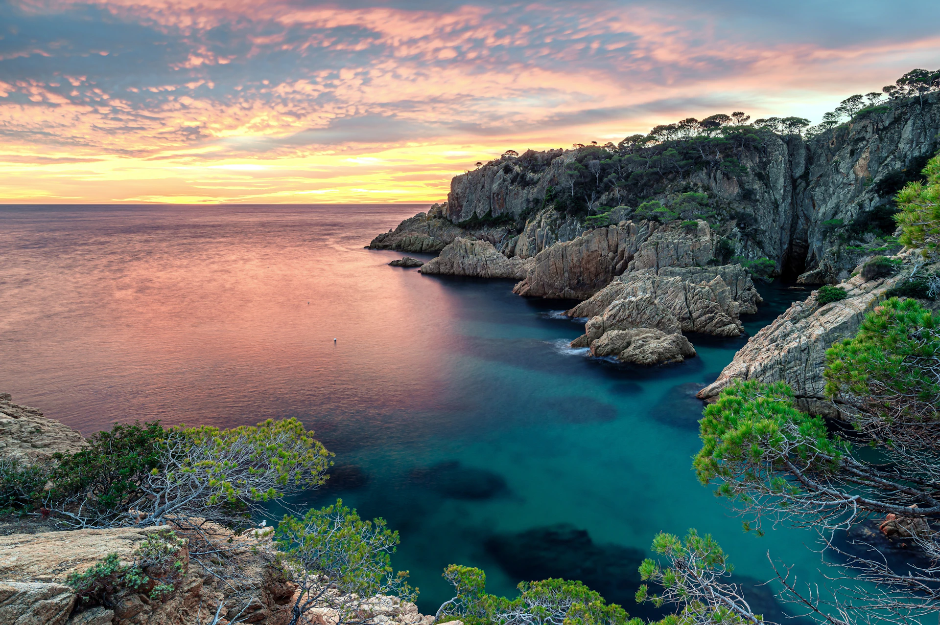 Rocky Mediterranean cove with turquoise water at sunset, cliffs and vegetation visible.