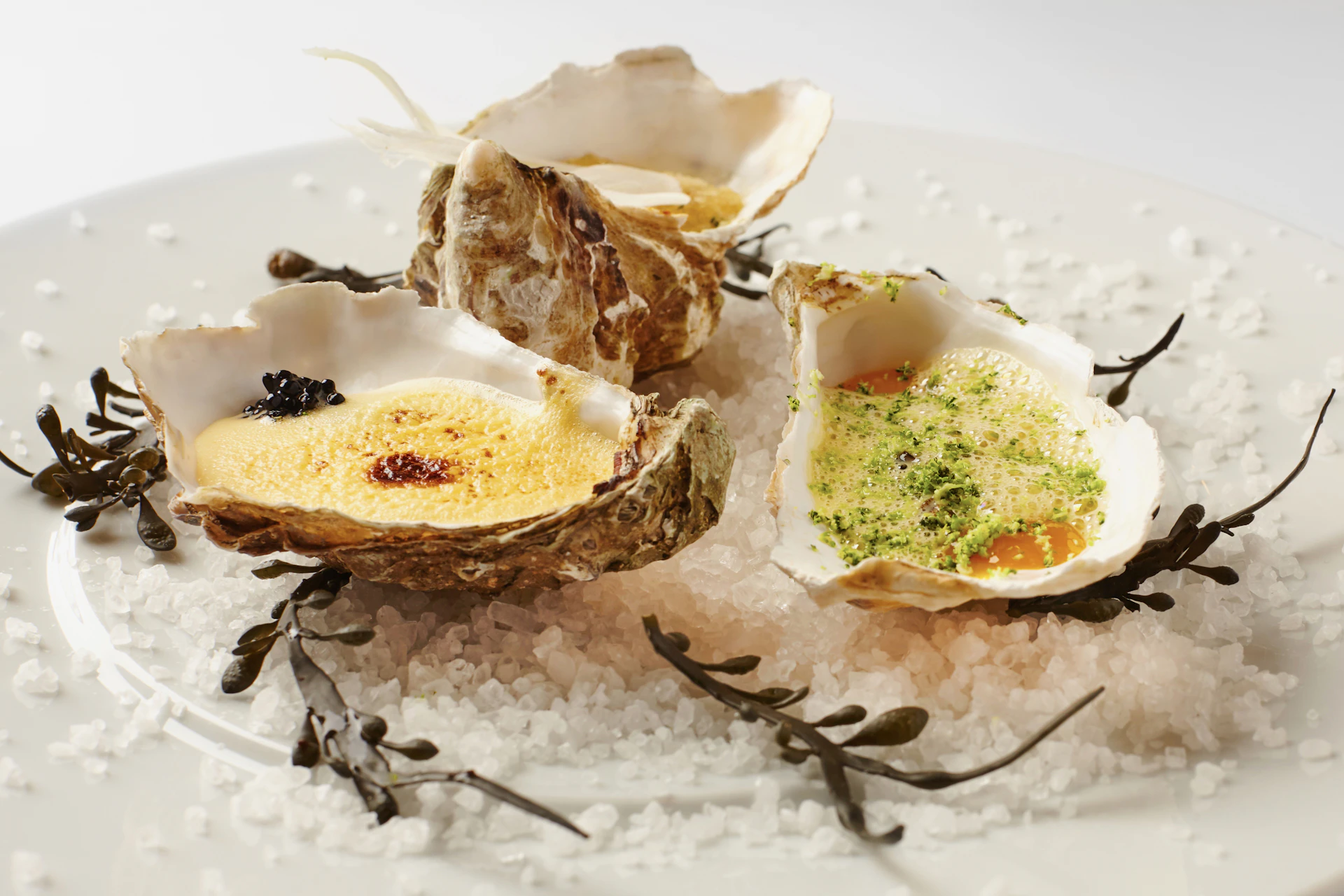 Three oysters on sea salt garnished with seaweed and topped with yellow and green preparations.