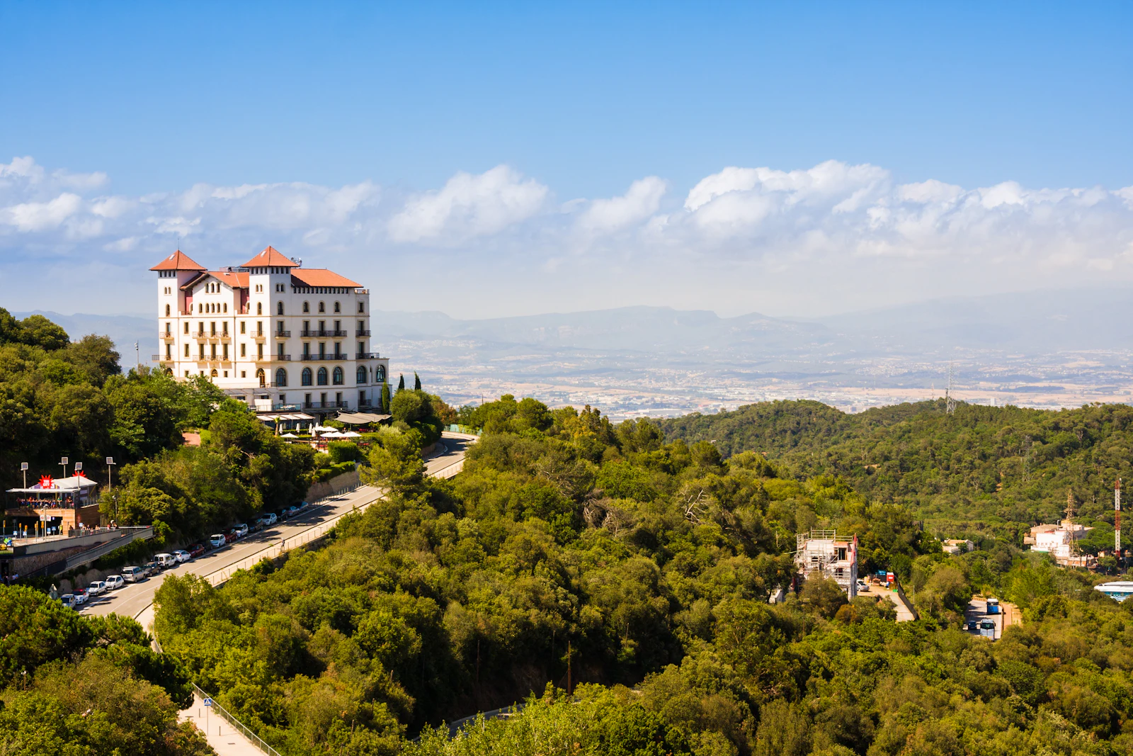 Large hilltop building with red roofs overlooking forested slopes and distant mountains.