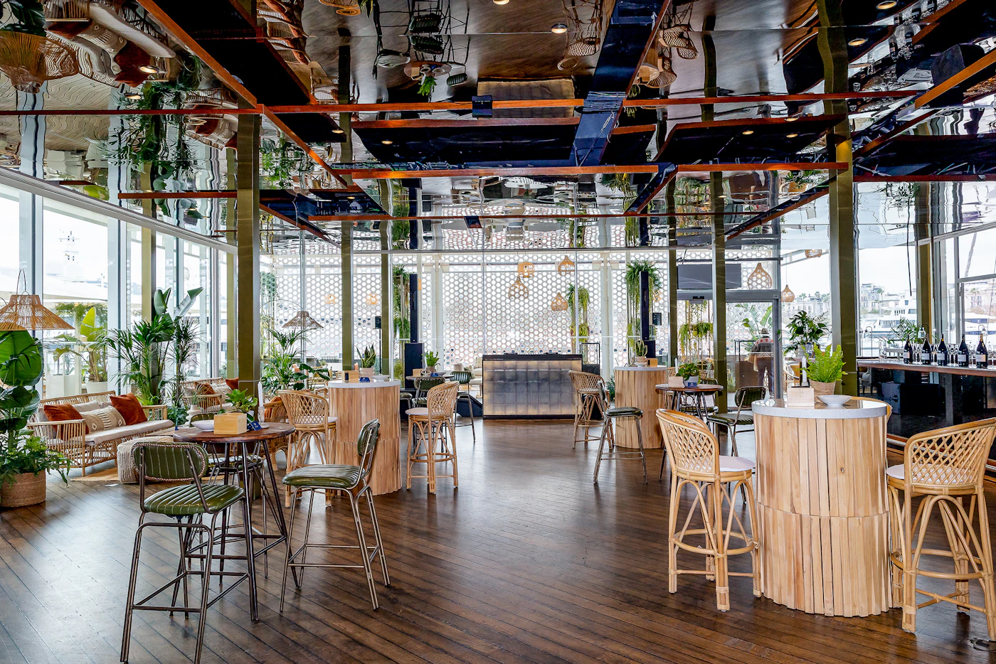 Bright bar interior with rattan high tables, stools, plants and a mirrored ceiling.