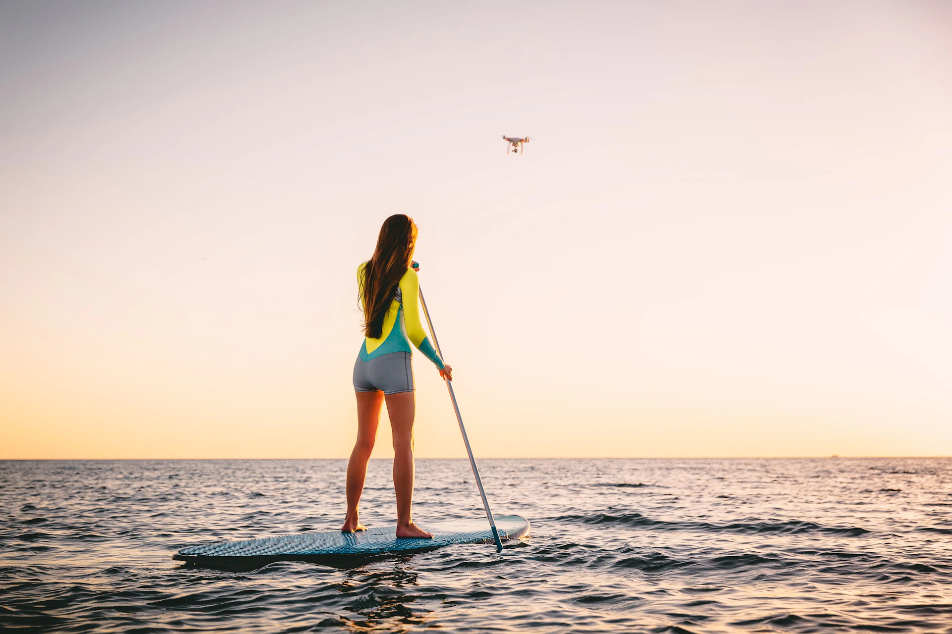 Mujer haciendo paddleboarding al atardecer mientras maneja un dron sobre su cabeza.