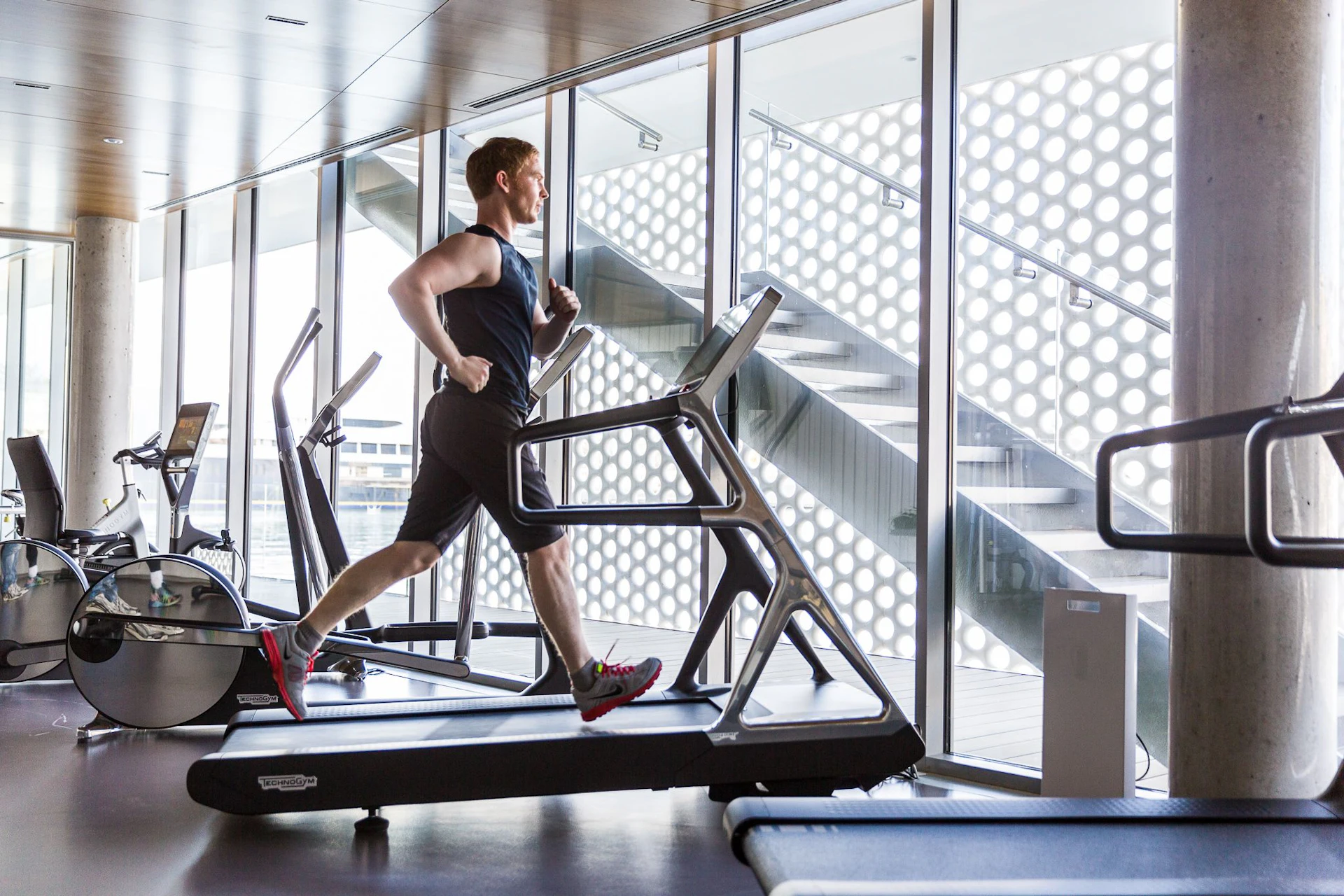 Man running on a treadmill in a bright gym with floor-to-ceiling windows overlooking Barcelona.