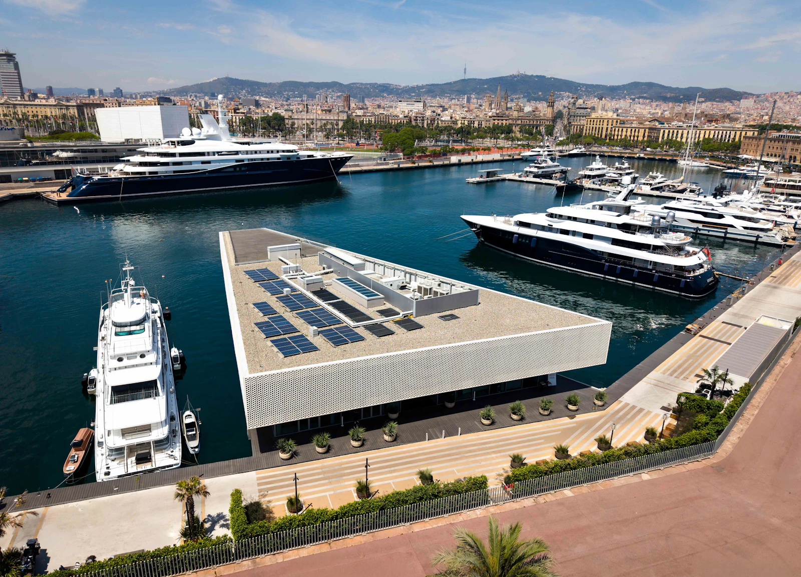 Aerial view of superyachts moored at a marina with Barcelona city skyline behind.