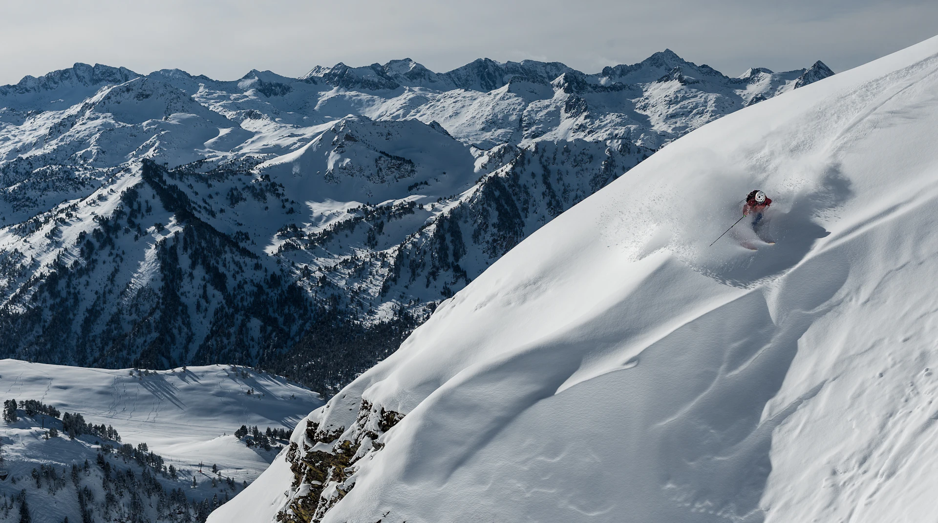 Skier descending a steep snowy mountain slope with snow-covered peaks in the background.